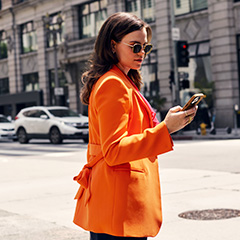 Woman in an orange jacket using her phone on a city street.