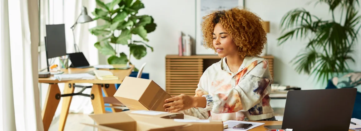 Person packing a small box at a desk with a laptop in a home workspace setting.