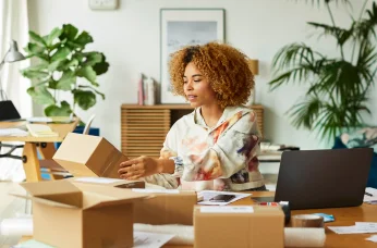Person packing a small box at a desk with a laptop in a home workspace setting.