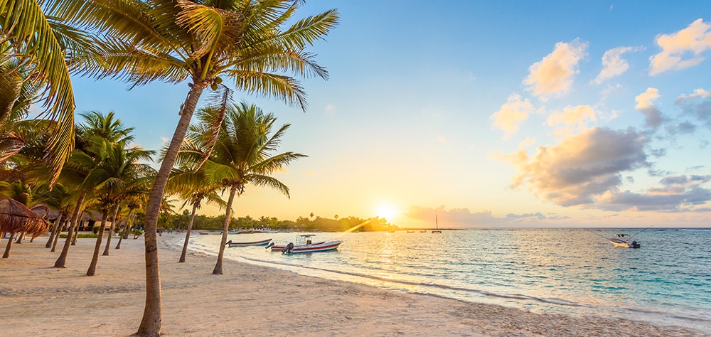 Sunrise over a palm lined beach in Cancun with boats floating on calm Caribbean waters. 