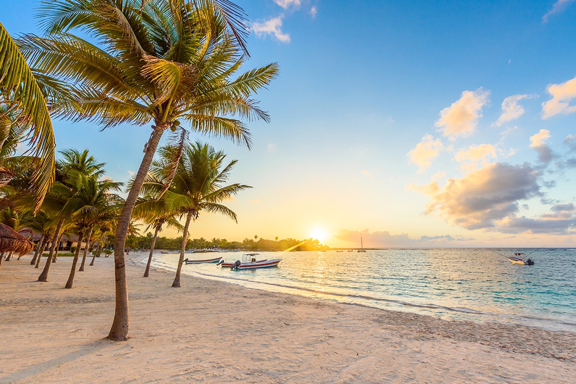 Sunrise over a palm lined beach in Cancun with boats floating on calm Caribbean waters. 