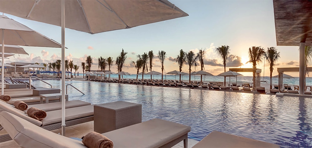 Luxury beachfront resort featuring poolside, loungers, umbrellas, palm trees, and ocean Sunset view. 