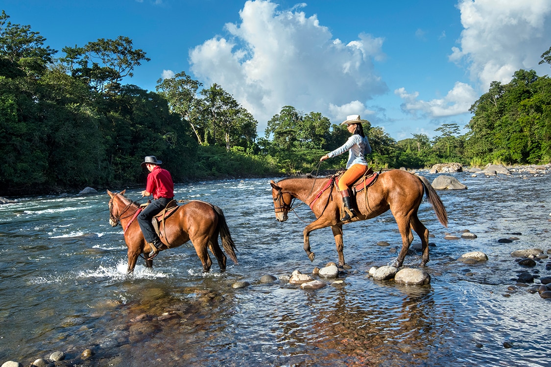 Travelers horseback riding across a river in Costa Rica surrounded by lush forest. 