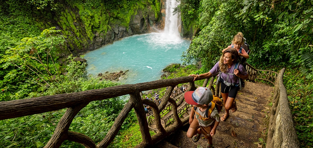 Family hiking along jungle trail beside turquoise waterfall pool in Costa Rica. 