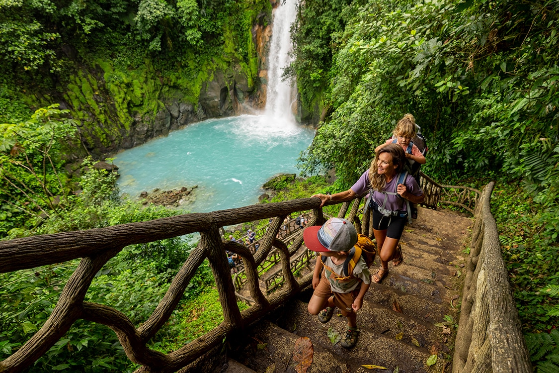 Family hiking along jungle trail beside turquoise waterfall pool in Costa Rica. 