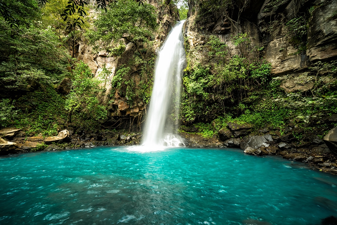 Waterfall cascading into a turquoise pool surrounded by lush tropical rainforest. 