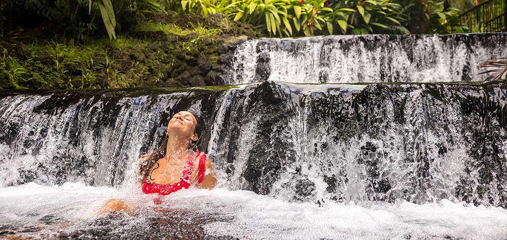 Woman relaxing under a natural waterfall in Costa Rica surrounded by lush tropical greenery. 