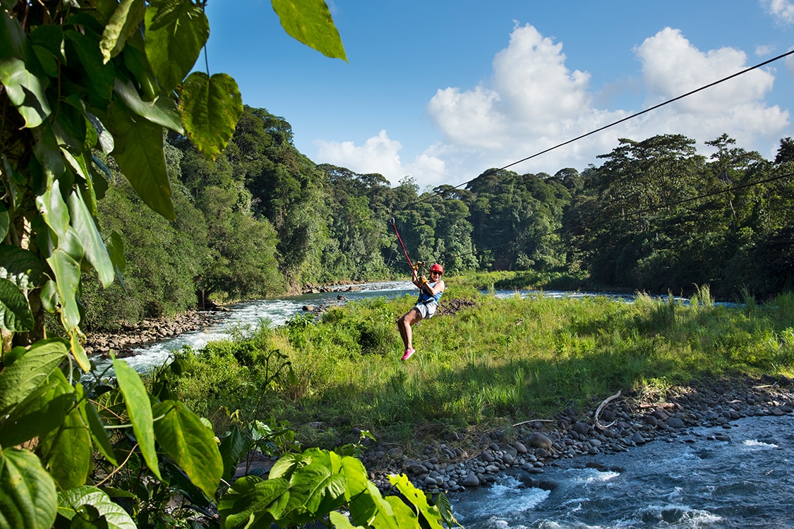 Woman zip lining over a lush river valley surrounded by tropical rainforest. 