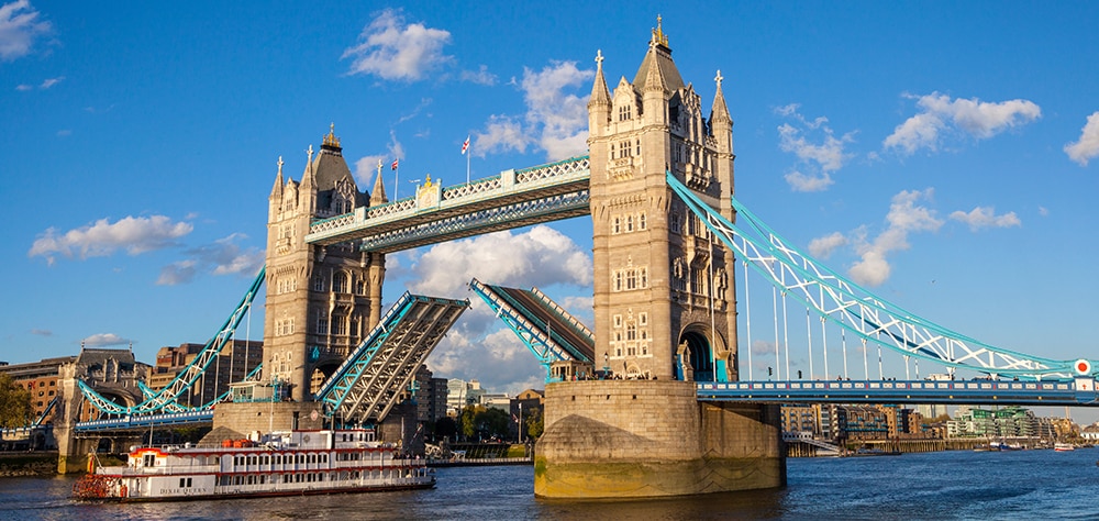 London's Tower Bridge opening over the River Thames with historic towers and blue suspension cables on a sunny day. 