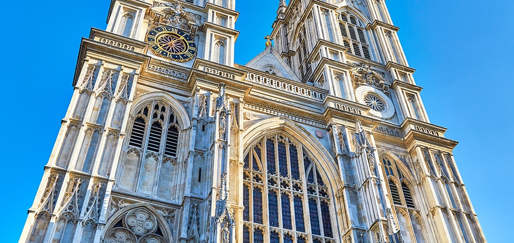 Historic Westminster Abbey in London featuring Gothic stonework, arched windows, and clock tower on a bright sunny day. 