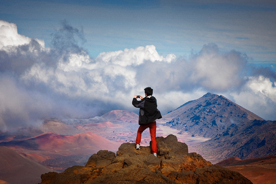 Traveler standing on rocky mountain summit photographing a dramatic volcanic landscape with mountains and clouds. 