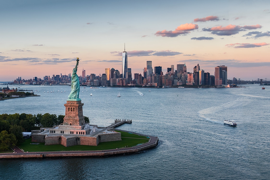 Statue of Liberty with Manhattan skyline and One World Trade Center at sunset over New York Harbor. 