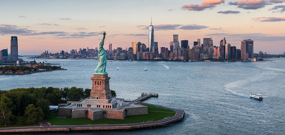 Statue of Liberty with Manhattan skyline and One World Trade Center at sunset over New York Harbor. 