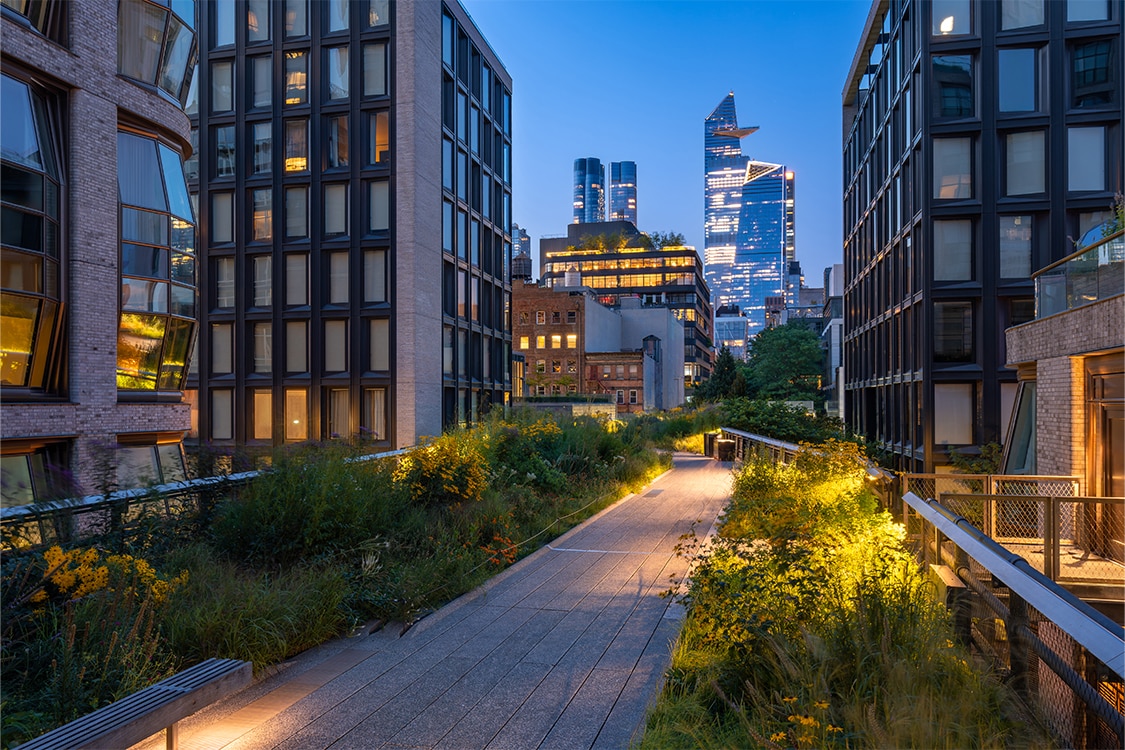The High Line at dusk with illuminated walkway and New York City skyline in the background. 