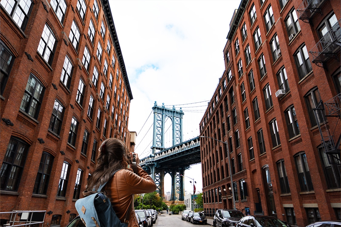 Tourist photographing the Manhattan Bridge in Dumbo Brooklyn between historic red brick buildings.