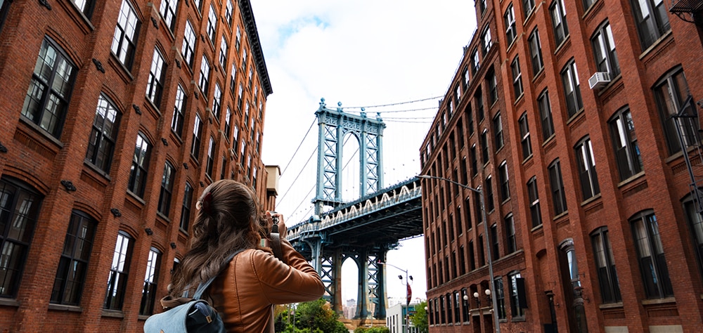 Tourist photographing the Manhattan Bridge in Dumbo Brooklyn between historic red brick buildings.