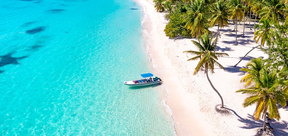 Boat anchored along a white sand beach with turquoise water and palm trees. 