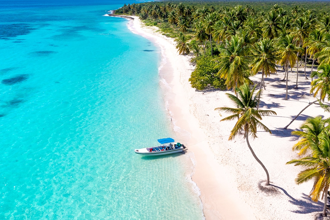 Boat anchored along a white sand beach with turquoise water and palm trees. 